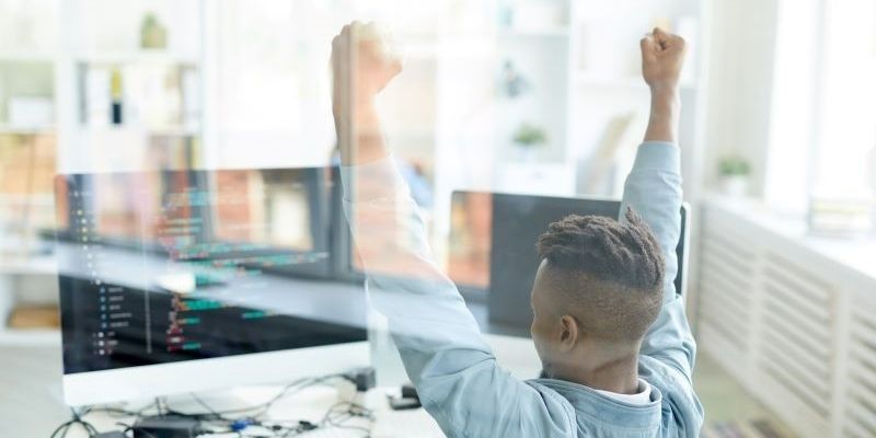 A young African-American AWS developer raises his arms in success in front of a coding terminal.